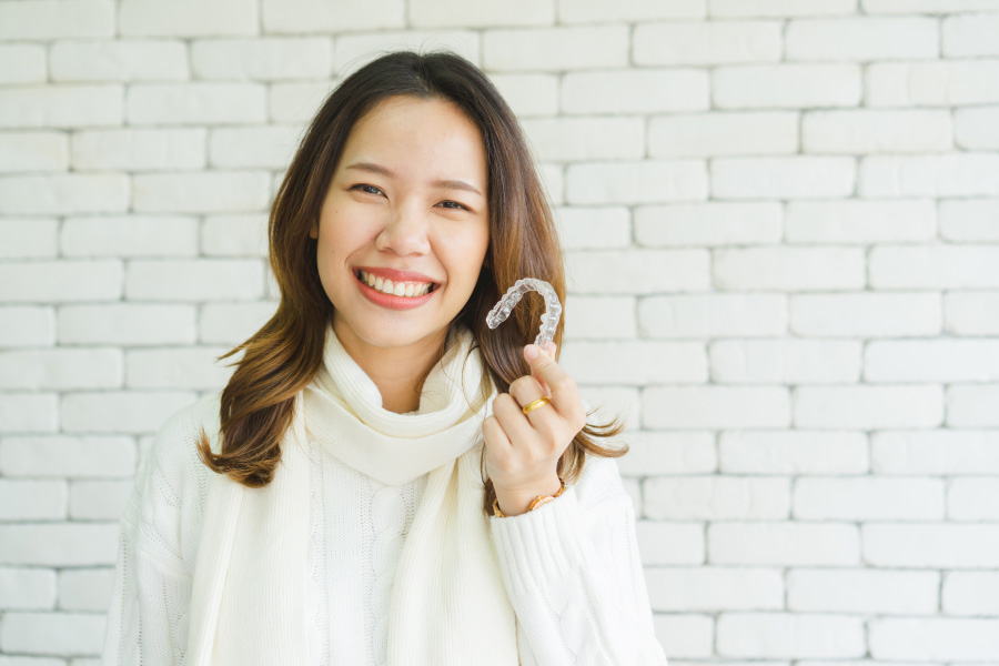 Asian woman in a cream turtleneck sweater smiles as she holds up her Invisalign aligners