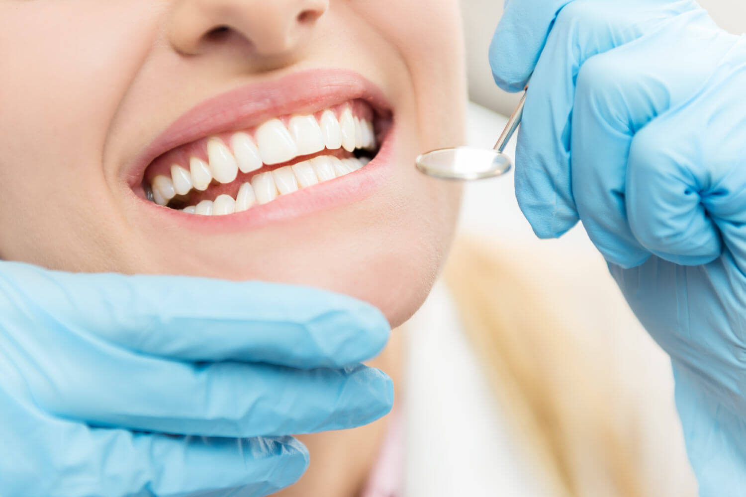 Close up of a woman's smile next to blue gloved hands holding a special dental mirror during her dental cleaning