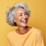 A gray-haired woman in a yellow blouse smiles brightly against a yellow background after getting restorative dentistry
